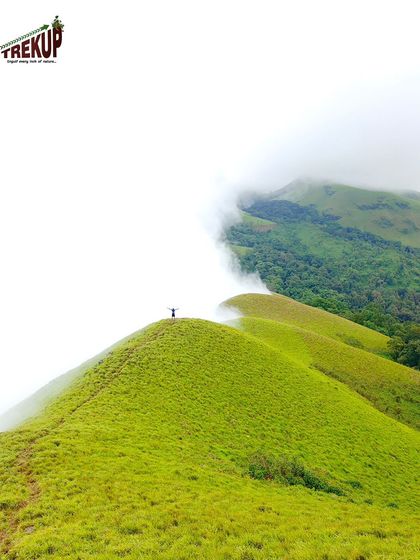A lone trekker stands on a green hill at Netravathi, dwarfed by the vastness of the landscape and the clouds.