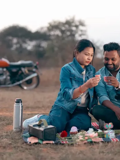 A picnic for two after a long ride. The couple enjoys a quiet moment together on a blanket, with their Royal Enfield parked in the background, a faithful companion on their journey.