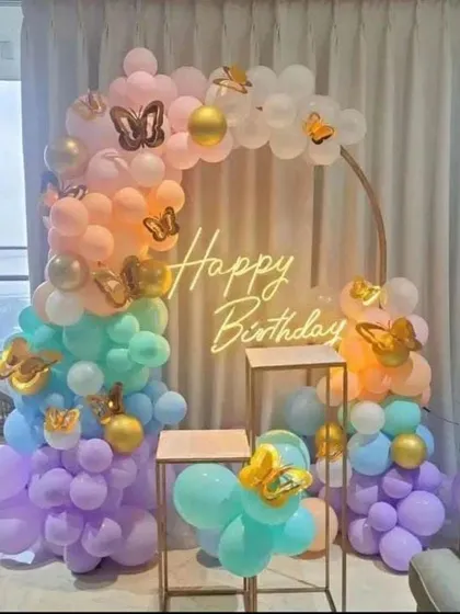 A rainbow pastel butterfly theme for a birthday party. This decoration features a circular balloon arch, a "Happy Birthday" neon sign, and gold butterfly accents.