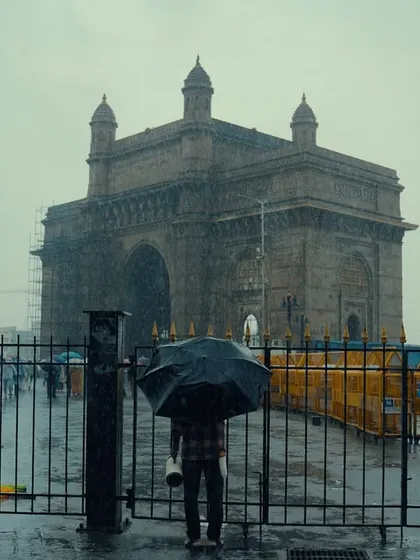 A solitary figure stands with an umbrella before the Gateway of India on a rainy day. The composition emphasizes a sense of scale and solitude, with the iconic monument appearing almost ethereal in the mist.