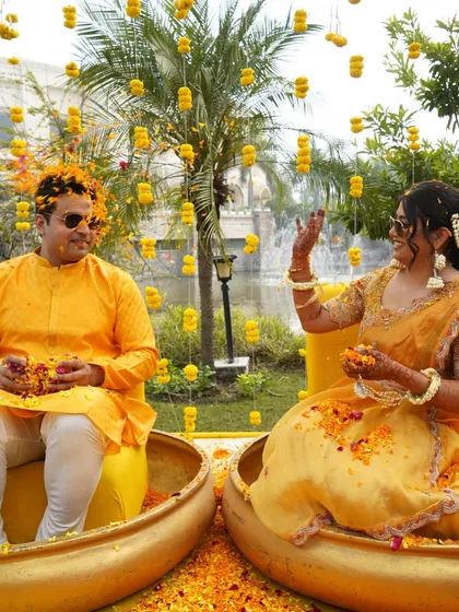 A playful moment during the Haldi ceremony, with the couple seated in large golden urlis, surrounded by a shower of flower petals.