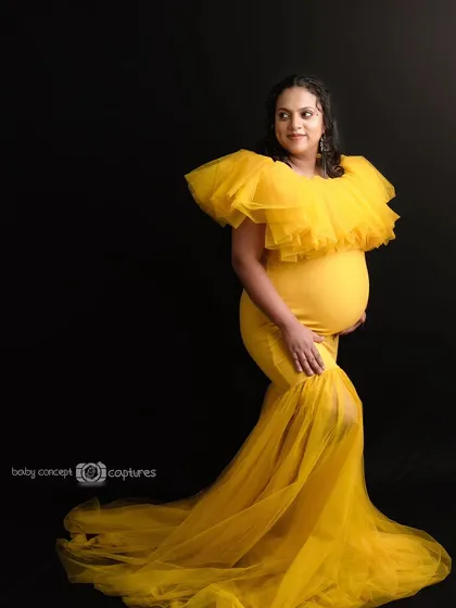 A full-length portrait of a woman showcasing a glamorous yellow ruffled maternity gown. The solid black background makes the color pop, creating a dramatic and fashionable studio shot.