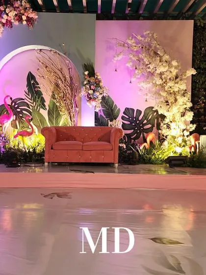 The main stage for a vibrant Mehendi ceremony. The setup includes a comfortable pink sofa for the bride, set against a backdrop of tropical leaves, pampas grass, and cherry blossoms.