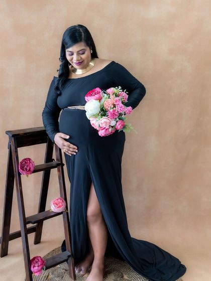 A chic and elegant studio portrait. The mom-to-be looks stunning in a black off-shoulder gown, holding a bouquet of pink and white flowers.