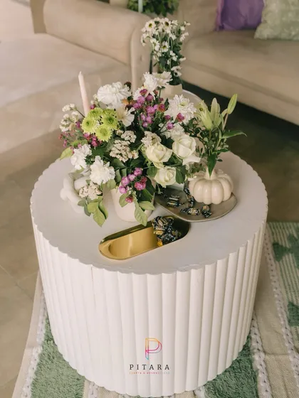 A modern centerpiece on a textured white table. The arrangement of fresh white and green florals is paired with a small white pumpkin prop and a gold accent dish, creating a chic and stylish look.