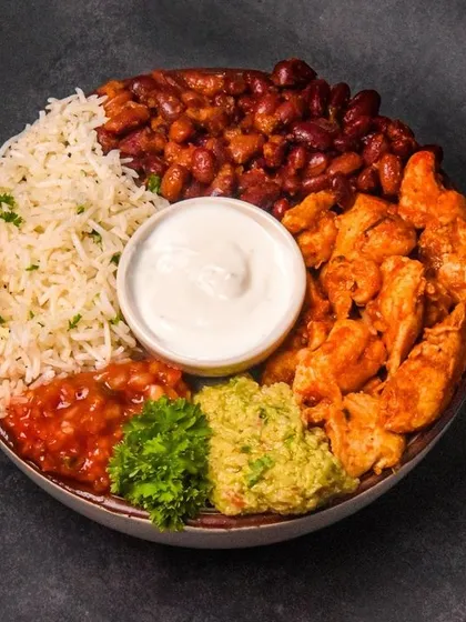 A vibrant and healthy chicken burrito bowl, neatly arranged with cilantro lime rice, seasoned chicken, kidney beans, fresh guacamole, and salsa. The top-down flatlay composition on a dark surface highlights each fresh ingredient.