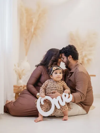 The birthday girl holds a "One" prop while her parents embrace lovingly behind her. This is a perfect family portrait to commemorate a first birthday milestone.