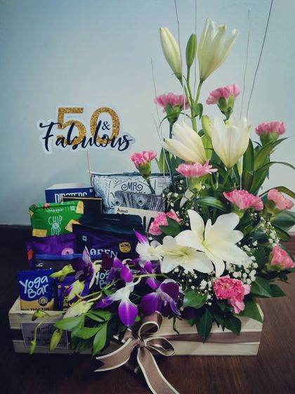 A beautiful and sophisticated 50th golden anniversary hamper. This gift combines a stunning arrangement of fresh lilies and carnations with a selection of healthy snacks and treats, topped with a '50 & Fabulous' sign.