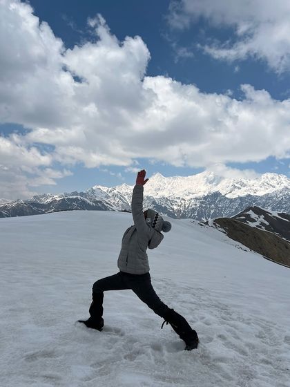 Holding Virabhadrasana I, or Warrior I, on the snow-covered peak. This pose embodies the strength and determination it takes to complete a high-altitude trek, feeling grounded and powerful against the elements.