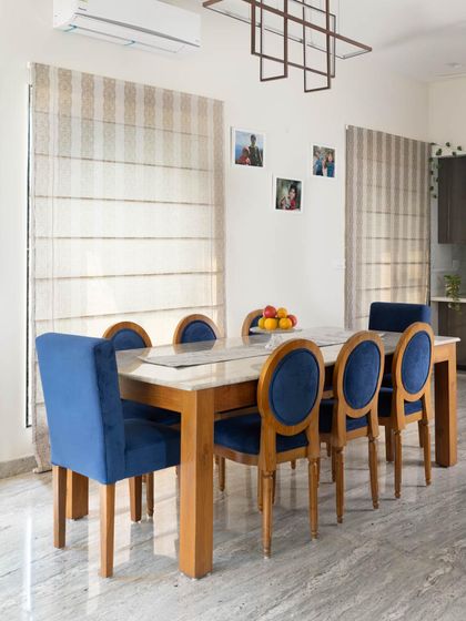 The dining area in the Lakshya villa, featuring a solid wood table and striking blue velvet chairs. Large windows with Roman blinds allow for plenty of light, making it a bright and inviting space for family meals.