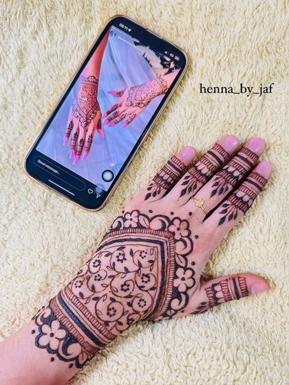 Recreating a client's requested design, this image shows the finished henna next to the inspiration photo on a phone. The pattern is a full coverage Gulf style design.