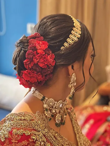 A classic bridal bun decorated with vibrant red carnations and a traditional golden matha patti. This is a bold and beautiful choice for a bride wearing a classic red lehenga, ensuring the hairstyle is as stunning as the outfit.