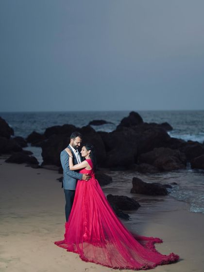 A dramatic pre-wedding photo on the beach at dusk. The long, flowing trail of the red gown creates a stunning silhouette against the evening sky.