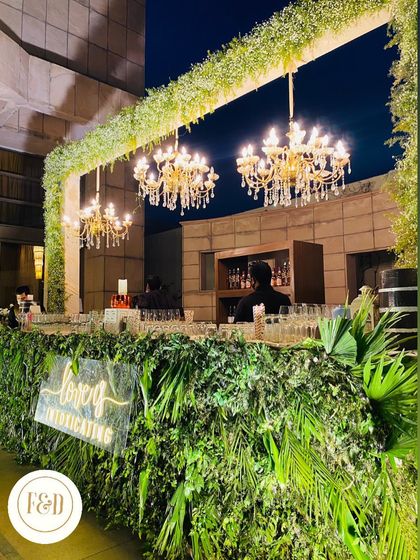 A lush and green bar design. The entire bar front is covered in a variety of ferns and tropical leaves, with elegant chandeliers hanging above for a touch of glamour.