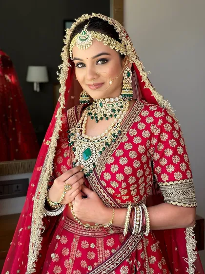 This bride looks stunning in her red lehenga, with makeup that enhances her features and complements her emerald jewelry. A perfect example of a classic bridal look.