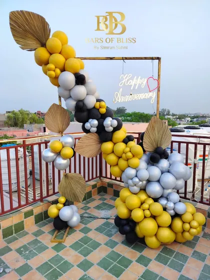 A stylish rooftop anniversary celebration. This modern setup features a simple frame decorated with a garland of yellow, black, and silver balloons, accented with dried palm leaves for a touch of boho chic.