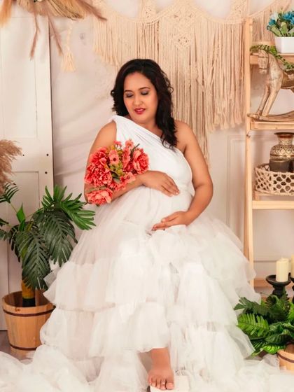 A serene shot of a mom-to-be in our white one-shoulder ruffled gown, holding a bouquet of flowers.