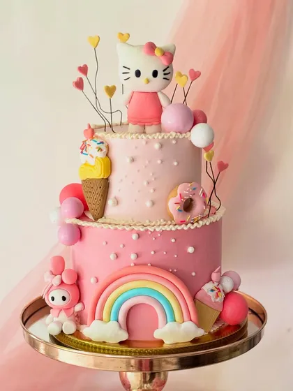A whimsical two-tier Hello Kitty cake designed for a magical birthday. The cake is adorned with edible rainbows, ice cream cones, donuts, and beloved Sanrio characters, all crafted from buttercream and sugar paste.