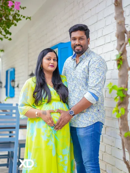 A sweet couple's portrait with hands forming a heart shape on the baby bump, set against a white brick wall.