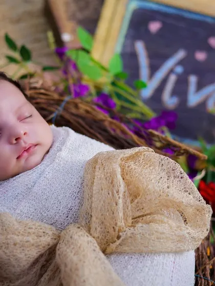 A newborn named Vivaan, swaddled in white and resting in a rustic nest adorned with purple flowers and a single red rose.