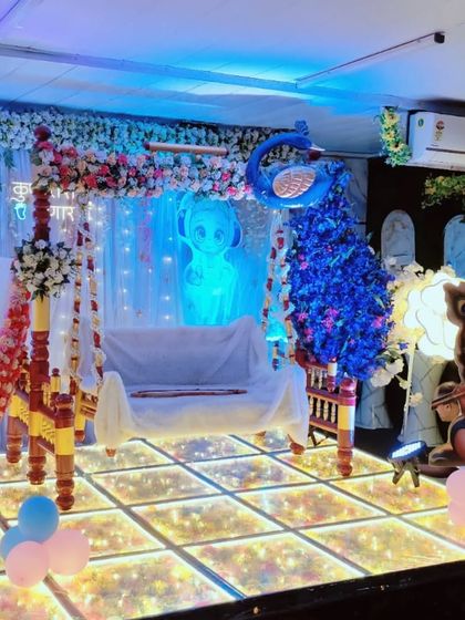 A traditional wooden swing (jhula) decorated for a naming ceremony with a Krishna theme. The LED floor and blue lighting create a modern yet spiritual feel for this important family function.