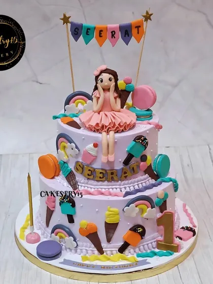 A sweet two-tier Candyland-themed cake for a first birthday. This chocolate cake is decorated with an adorable fondant doll, macarons, ice cream cones, and other candy elements.