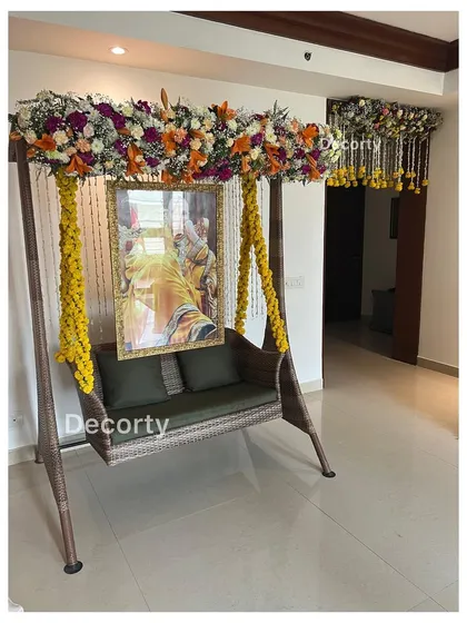 A beautifully decorated swing (jhoola) for a Mundan ceremony, adorned with yellow floral strings and a lush overhead flower arrangement. A portrait of a deity hangs in the background, adding a spiritual touch.