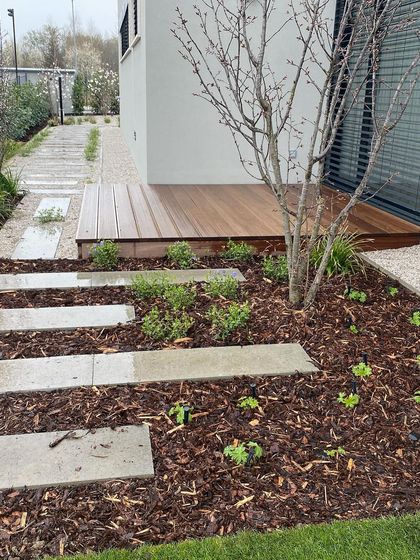 A work-in-progress shot of a modern garden. The design includes a wooden deck, gravel paths, and concrete stepping stones laid over bark mulch, showing the foundational layers of the landscape.