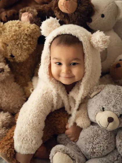 A sweet baby in a teddy bear hoodie peeks out from a pile of stuffed animals. This is one of my favorite setups for a sitter session.