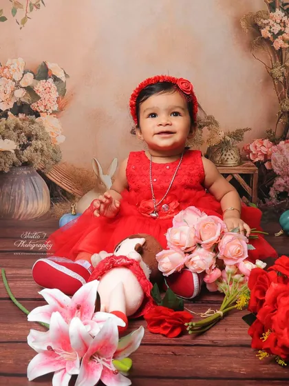 This little girl is surrounded by a garden of flowers in this studio setup. The rich colors of her red dress and the soft floral background create a warm and joyful baby portrait.