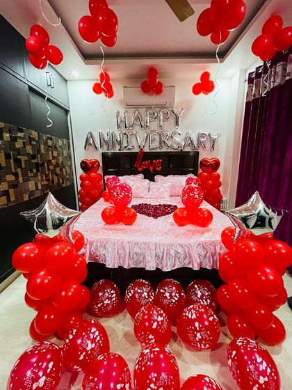 This is a classic all-red anniversary bedroom decoration. The setup includes "Happy Anniversary" foil letters, heart-shaped balloon arrangements, and a bed decorated with rose petals for a passionate and romantic surprise.