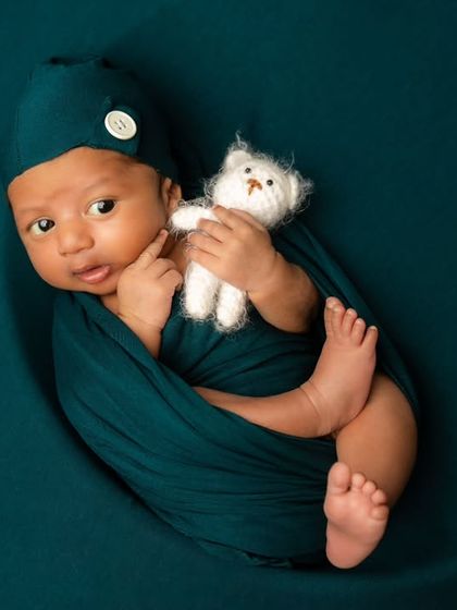 Wide-eyed and wonderful. This awake newborn is nestled in a moon prop, wrapped in a deep teal swaddle and holding a tiny teddy bear, looking right at the camera.
