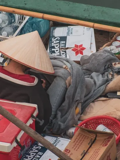 A close-up shot of a person in a conical hat resting in a small boat filled with goods, offering a glimpse into the life of a floating vendor.