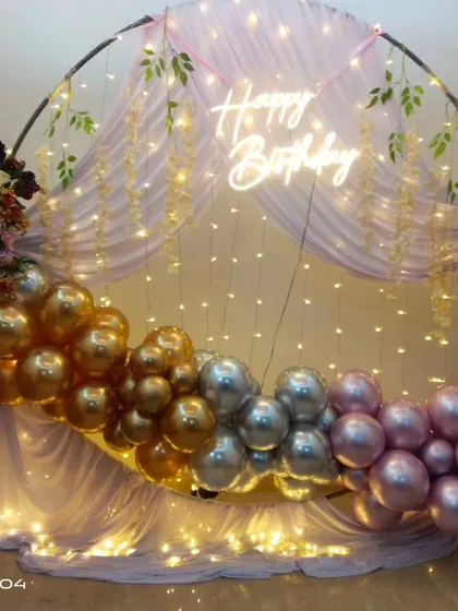 A beautiful birthday backdrop with a round frame, sheer drapes, fairy lights, and a garland of chrome gold, silver, and pink balloons.