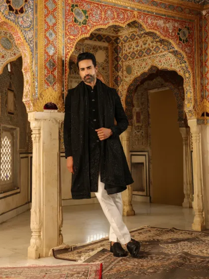 The beauty of our craft lies in the details. This image shows a model in a stunning black Indo-Western sherwani with a matching stole, set against a backdrop of ornate architecture that complements the outfit's traditional elegance.
