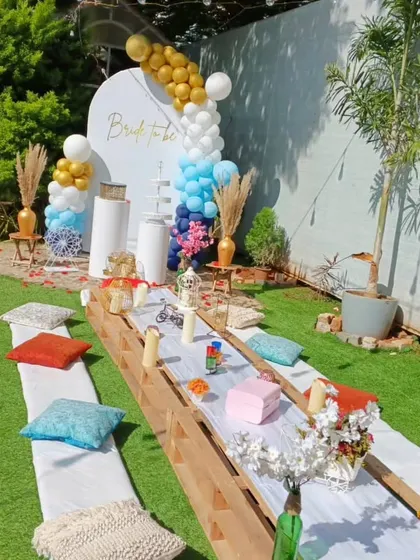 A bright and sunny bachelorette picnic. The long pallet table is decorated with colorful cushions and a stylish blue, white, and gold balloon backdrop for a cheerful daytime celebration.
