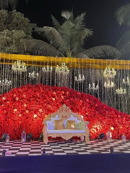 A bold and dramatic reception stage featuring a massive wall of red roses. The design is accented with hanging chandeliers and a checkered floor, creating a look of pure opulence.
