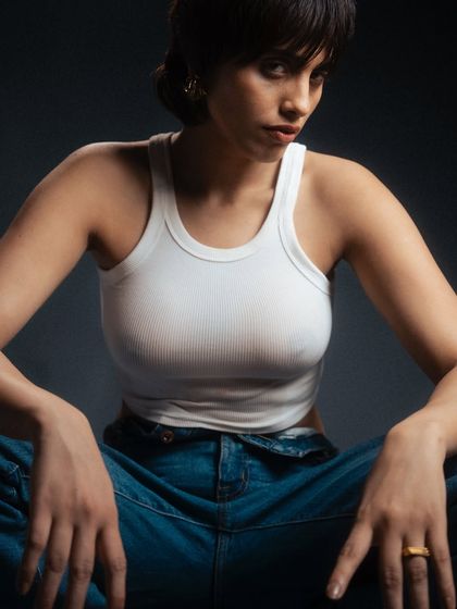 A close-up studio portrait with a moody, dark grey background. The lighting is sculpted to emphasize the model's features and the texture of the white tank top, creating an intense and intimate feel.