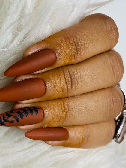 This image, shown on a practice hand, features a matte brown stiletto nail with freehand black leaf art. You will learn how to work with matte top coats and create detailed art on different nail shapes.