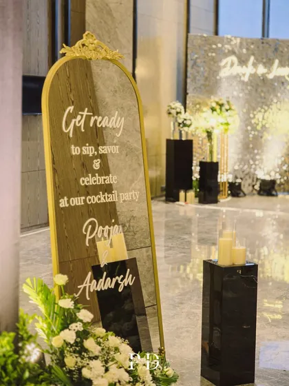 A chic welcome sign for a cocktail party, using a gold-framed mirror and elegant script. It perfectly complements the glamorous 'Drunk in Love' photo booth in the background.
