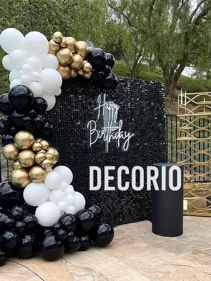 An outdoor poolside party setup with a black sequin wall as the centerpiece. The wall is framed by a classic black, white, and gold balloon arrangement for a touch of Hollywood glam.