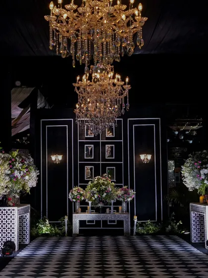 A breathtaking entrance for a monochrome-themed Sangeet. A checkered dance floor leads to a stage framed by massive floral arrangements and magnificent crystal chandeliers, setting a tone of pure luxury.