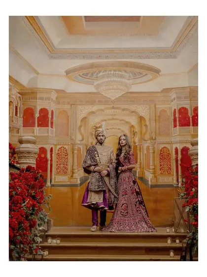 A portrait of pure royalty. The couple stands on the steps of a grand staircase in a Mumbai palace, their regal attire perfectly complementing the opulent surroundings.