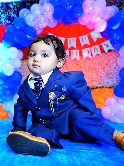 A full-length portrait of a smartly dressed toddler posing confidently on a blue mat during his birthday party photoshoot.