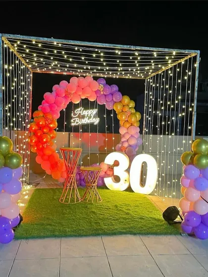A full view of a 30th birthday decoration on a rooftop. The setup includes a colorful circular balloon arch, a neon sign, and light-up marquee numbers, all framed within a structure of string lights.