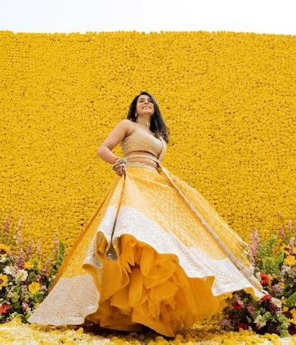A happy bride at her Haldi ceremony, posing in front of a vibrant yellow floral wall. The decor perfectly complements her attire.