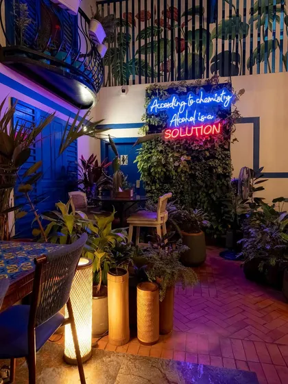 The Greek-themed courtyard transforms at night, with neon signs and ambient lighting creating a cool, vibrant atmosphere. The "Alcohol is a solution" sign adds a touch of humor and becomes a focal point.