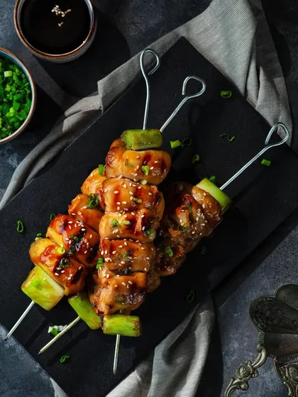 A top-down view of chicken skewers from The Khyber Resort's oriental menu. The dark slate plate and grey linen provide a rustic yet elegant backdrop, allowing the colors of the grilled chicken and garnishes to stand out.