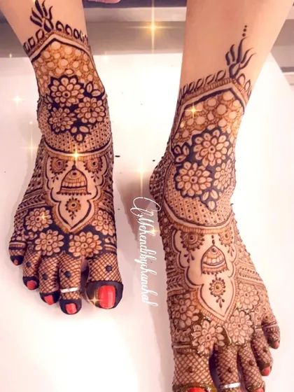 A close-up of intricate bridal feet mehendi, with a central bell motif surrounded by dense floral patterns covering the entire foot.