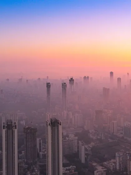 A panoramic drone shot of Mumbai's skyline enveloped in a thick layer of pink and yellow haze at sunset.